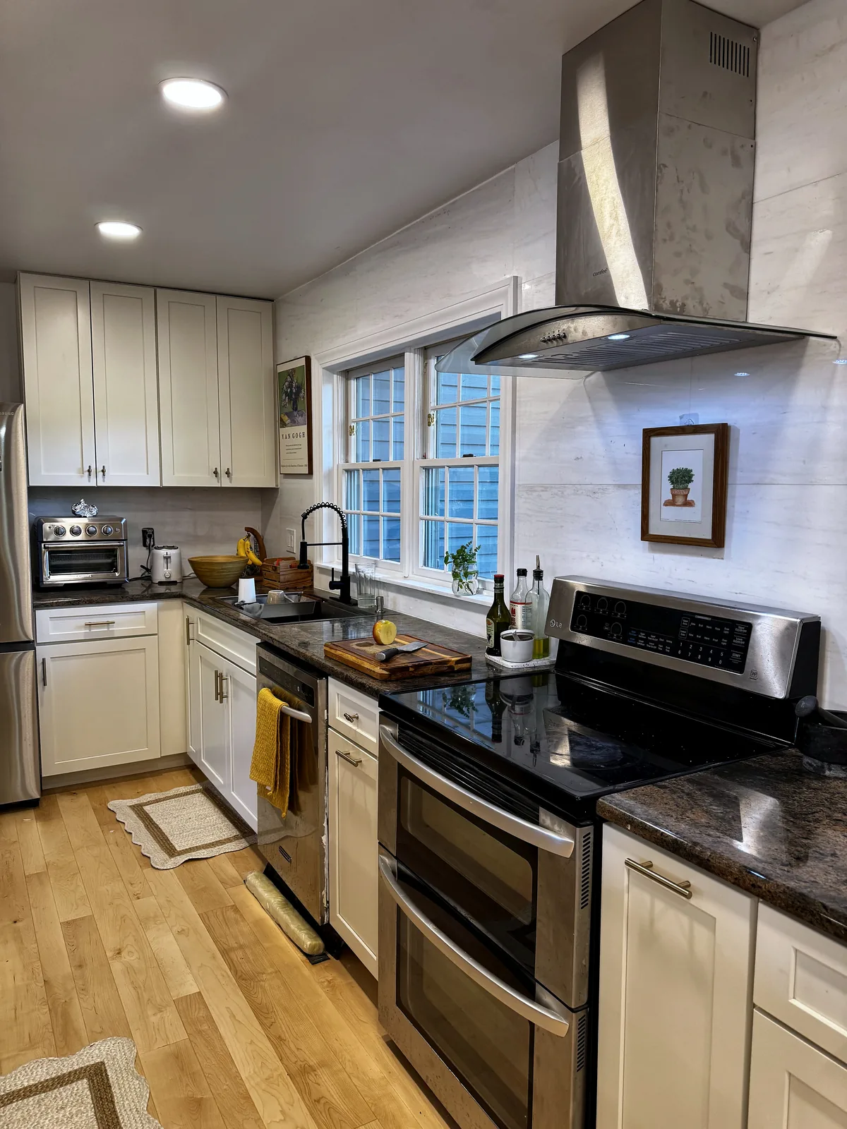 Finished kitchen range wall with white cabinets, stainless hood, window, and hardwood flooring